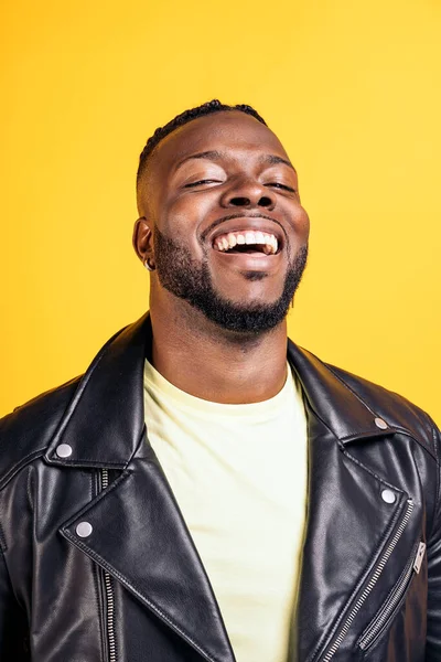 Happy black man wearing black leather jacket laughing in studio shot against yellow background.
