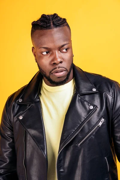 African american man with cool hairstyle and wearing black leather jacket posing in studio shot against yellow background.