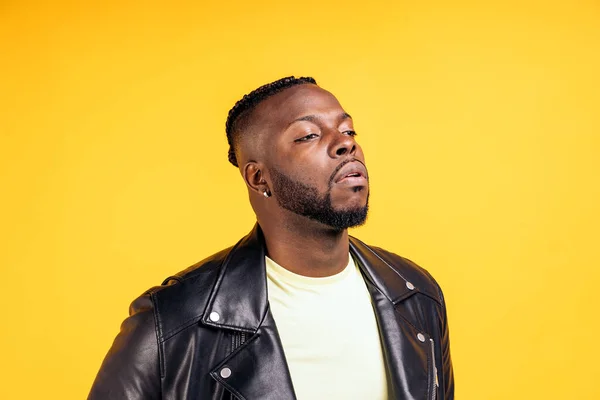 African american man with cool hairstyle and wearing black leather jacket posing in studio shot against yellow background.