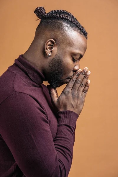 African american man with closed eyes and with his hands together posing in studio shot against brown background.