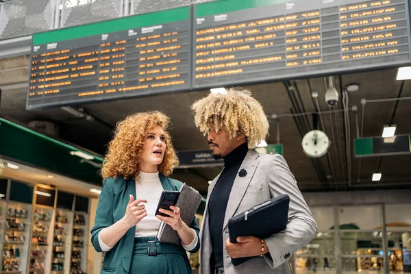 Business woman with formal wear talking with her african coworker and using mobile phone. They are in a train station in Madrid.