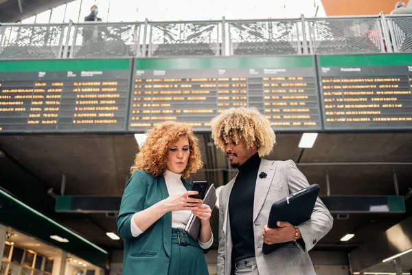 Business woman with formal wear talking with her african coworker and using mobile phone. They are in a train station in Madrid.