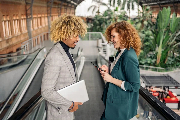 Business woman with formal wear using her mobile phone and talking with her african coworker in train station.