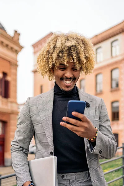 Happy black business man with afro hair and cool suit using his mobile phone in the city.