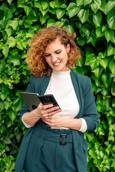 Happy business woman with curly hair and beautiful suit using her phone against green background with plants.