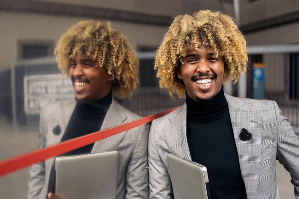 Happy black business man with afro hair and cool suit smiling and looking at camera outdoors. He is holding a laptop.