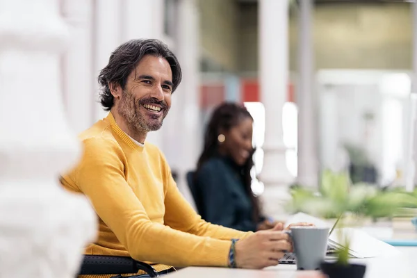 Bearded office worker sitting in the desk smiling and using his laptop. There is a black business woman in the background.
