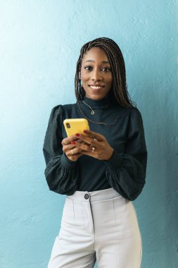 Smiley african office worker holding her mobile phone and looking at camera against blue background.