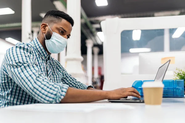 Focused black man with face mask sitting in his desk and using his laptop. Office concept.