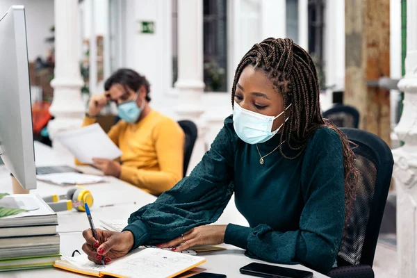 Focused black woman with face mask sitting in her desk and doing paper work next to other coworkers. Office concept.