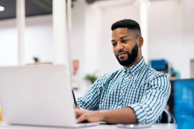 Afro american office worker sitting in his desk and using his laptop. He is smiling and looking the screen.