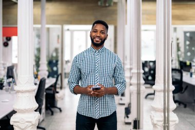 Smiley african office worker holding his mobile phone and looking at camera. Co-working concept.