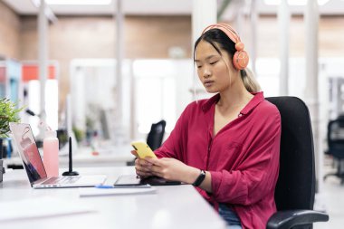 Focused asian woman with headphones sitting in her desk in the office and using her phone.