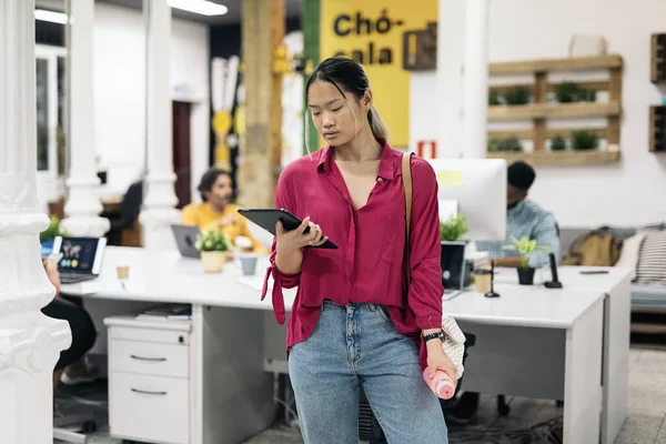 Young asian office worker standing in the office using her digital tablet holding a water bottle.