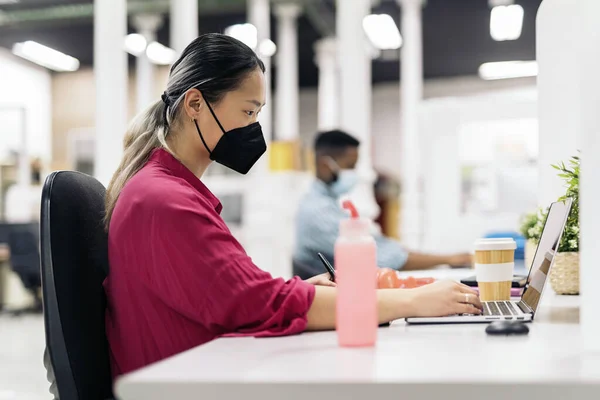 Concentrated asian office worker with face mask sitting in her desk and working with a laptop. Co-working concept.