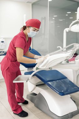 Female dentist working in modern dental clinic and wearing face mask. She is using spray with sanitizer to clean the equipment.