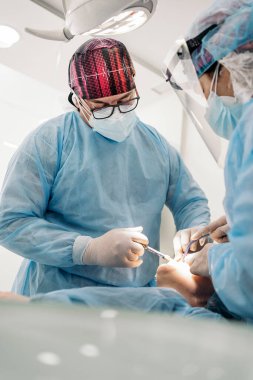 Male dentist and his team wearing face mask and protective clothes doing treatment to unrecognized patient.