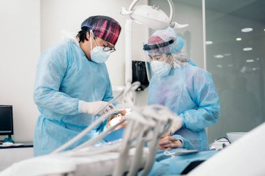 Male dentist and his team wearing face mask and protective clothes doing treatment to unrecognized patient.