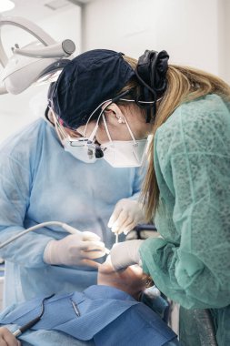 Female dentist wearing face mask and dental magnifying glasses doing treatment to unrecognized patient.