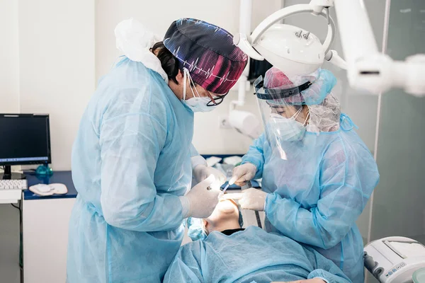 Male dentist and his team wearing face mask and protective clothes doing treatment to unrecognized patient.