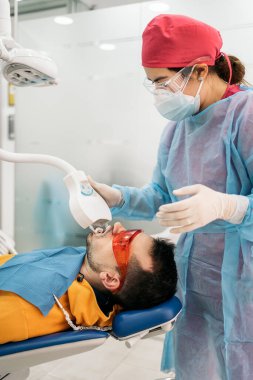 Man lying down in dental chair and wearing protective glasses using tooth whitening machine.