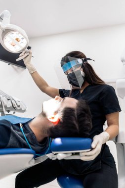 Female dentist wearing face shield isolation mask using light and working in modern clinic.