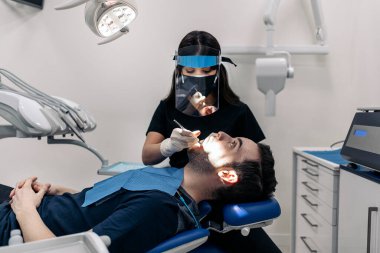 Female dentist wearing protective face mask working with young man during checkup revision.
