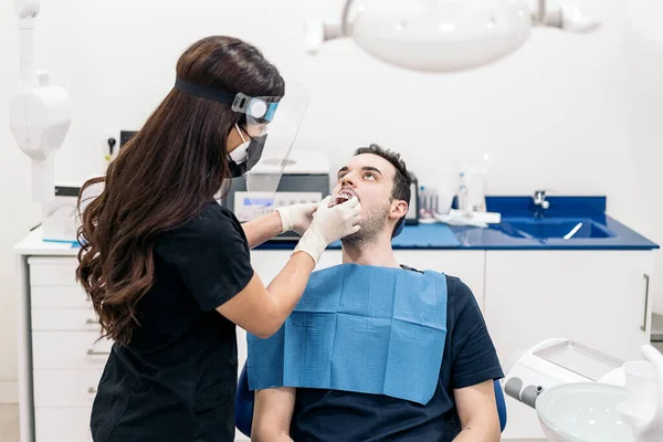 Female dentist wearing face shield isolation mask working with male patient and putting teeth covers.