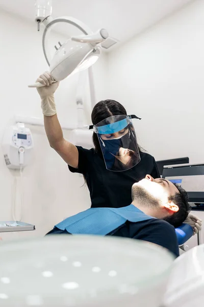 Female dentist wearing face shield isolation mask using light and working in modern clinic.