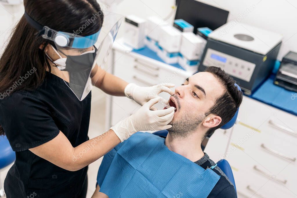 Female dentist wearing face shield isolation mask working with male patient and putting teeth covers.