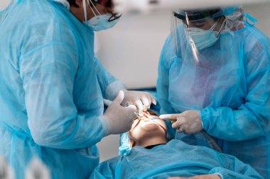 Dentist team wearing face masks and hair nets working in dental clinic with adult patient.