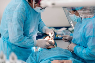 Dentist team wearing face masks and hair nets working in dental clinic with adult patient.