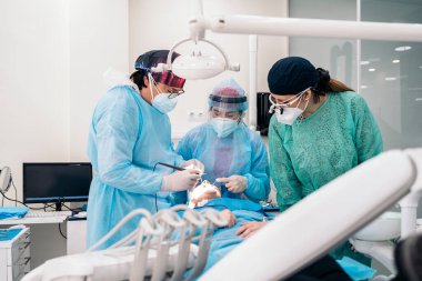 Dentist team wearing face masks and hair nets working in dental clinic with adult patient.