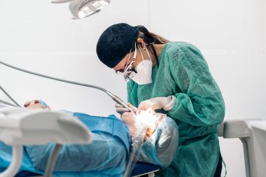 Dentist wearing face mask and dental magnifying glasses doing treatment to female patient.
