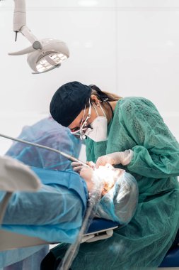 Female dentist wearing face mask and dental magnifying glasses doing treatment to unrecognized patient.
