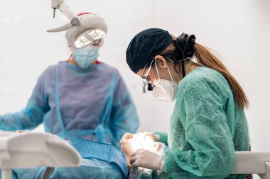 Female dentist wearing face mask and dental magnifying glasses doing treatment to unrecognized patient.