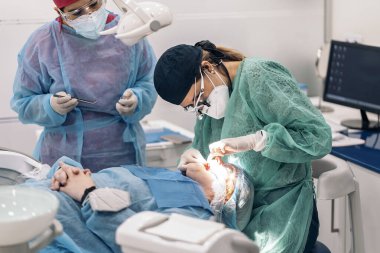Dentist wearing face mask and dental magnifying glasses doing treatment to female patient.