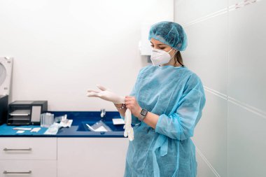 Female medical worker wearing face mask and protective clothes putting on latex gloves.
