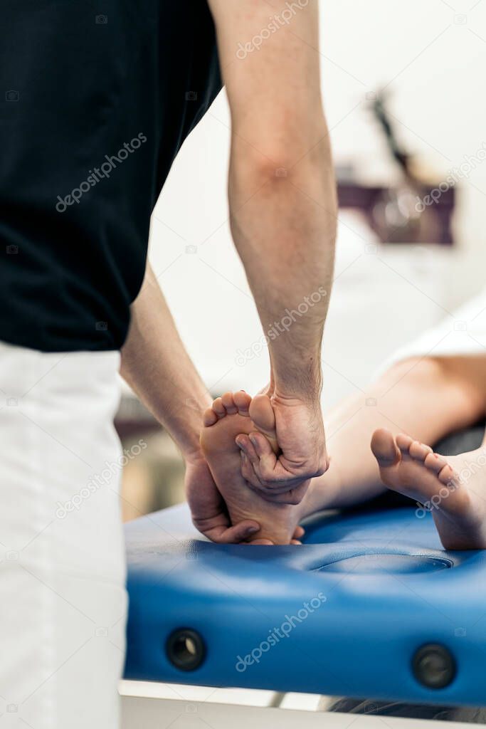 Male physiotherapist giving feet massage to unrecognized woman lying in stretcher.