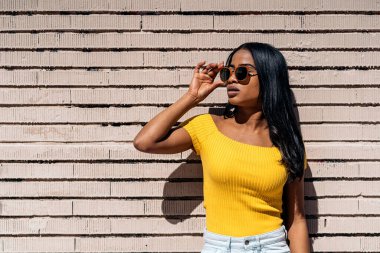 Güneş gözlüğü takan güzel ve kendinden emin genç bir kadın duvara bakıp poz veriyor..