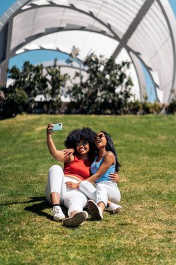 Güzel Afrikalı Amerikalı arkadaşlar cep telefonlarıyla selfie çekiyorlar ve güneşli bir günde parkta eğleniyorlar..