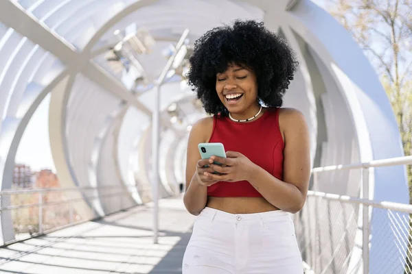 Cool young black woman with afro hair smiling and using mobile phone in the street.