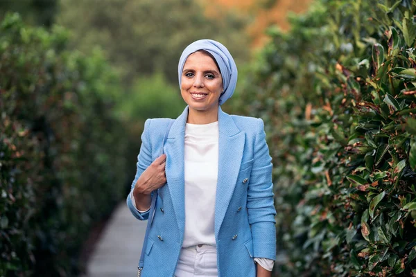 Front view of a smiling muslim woman standing among large bushes wearing a blue light suit and headscarf holding her purse on a pathway looking at camera in a sunny day.