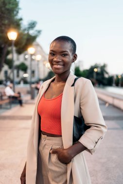 Şık, Afro-Amerikan, genç, yetişkin bir kadının gün batımında şehirde dikilerek gülüşü.