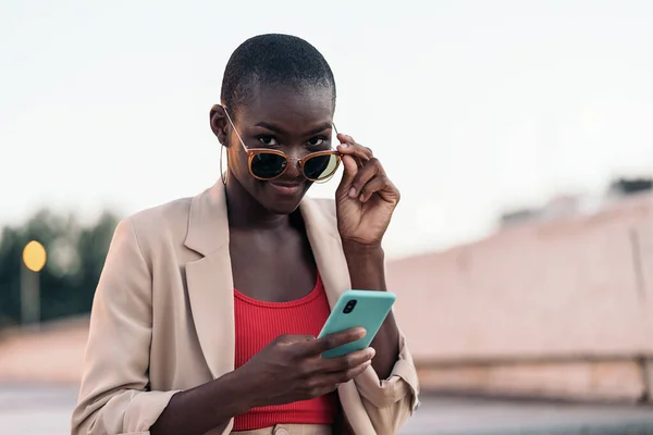 Elegant african american young adult woman holding sunglasses and using phone while looking at camera in an urban scene