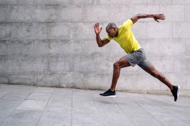 Sarı tişörtlü ve şortlu Afro-Amerikan bir sporcunun yan görüntüsü eğimli zemine doğru hızla koşuyor ve beton duvarın arkasında....