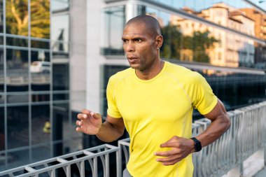 Sarı tişörtlü ve şortlu Afro-Amerikan bir sporcunun açık havada kaldırımda koşarkenki görüntüsü..