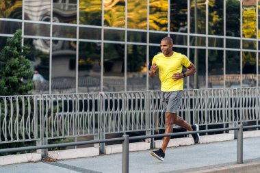 Sarı tişörtlü ve şortlu neşeli bir Afro-Amerikan sporcunun şehir dışında kaldırımda koşuşunun yan görüntüsü..