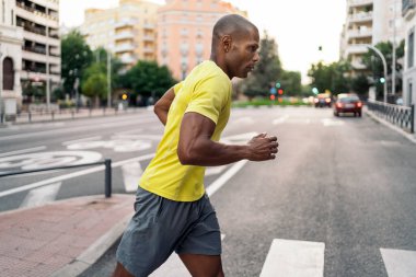 Sarı tişörtlü ve şortlu, güneşli bir günde yaya geçidinde koşan Afro-Amerikalı bir sporcunun yan görüntüsü..