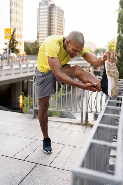 Front side view of African American man stretching his left leg over railing in the city,